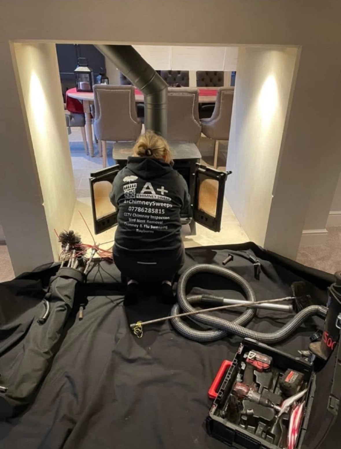 A+ Chimney Sweeps technician in branded uniform sweeping a stove with rods, vacuum and dust sheets in place